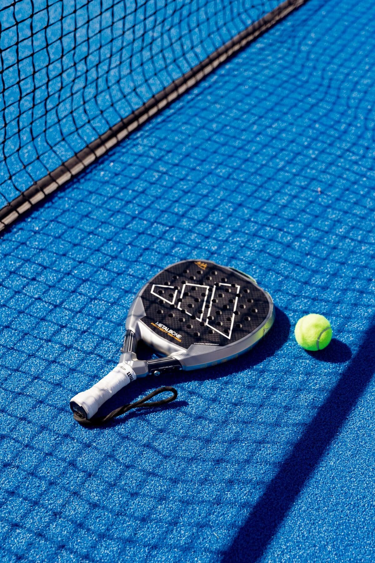 Una raqueta de pádel y una pelota sobre el piso azul de la cancha.